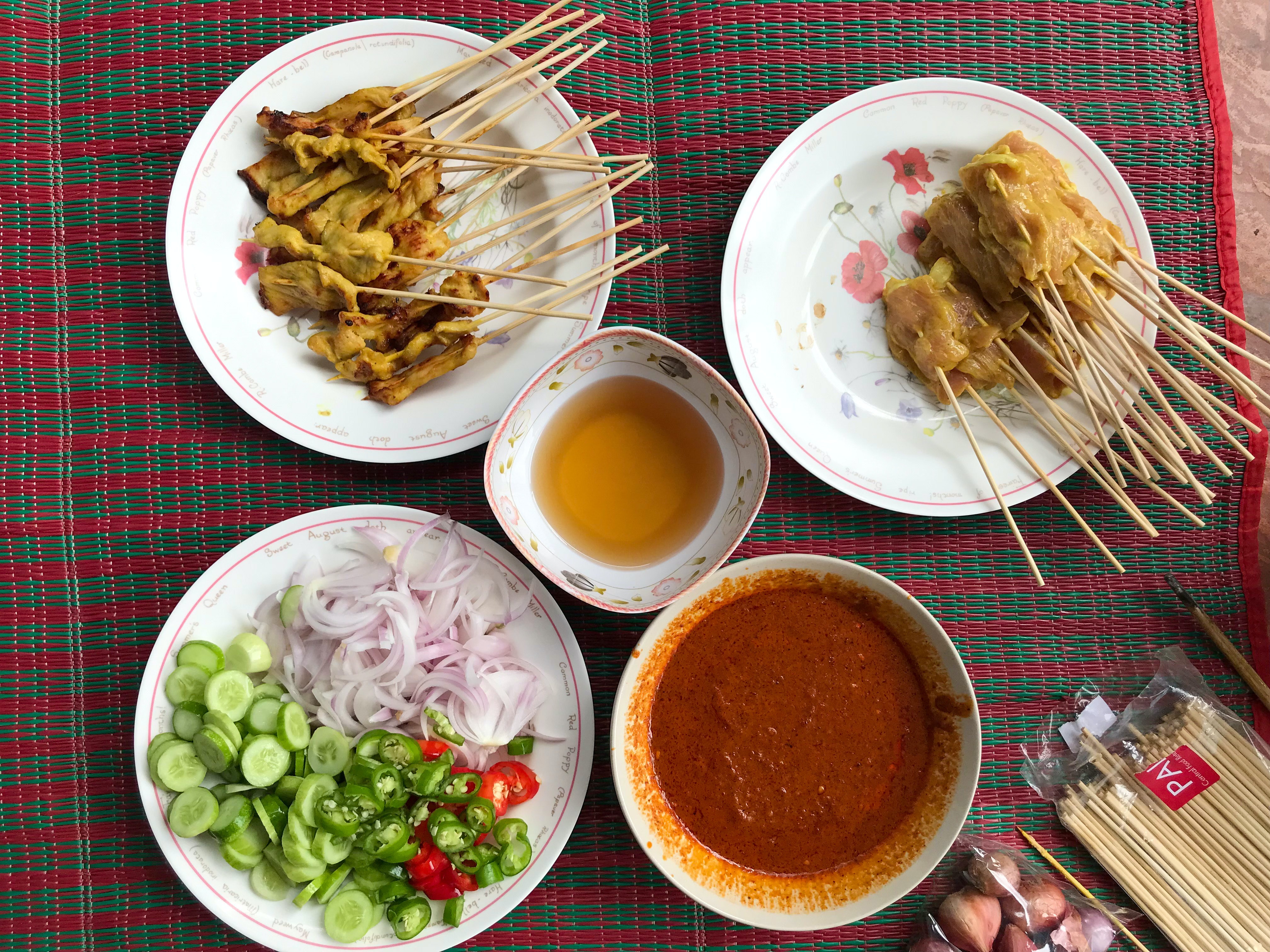 Two plates of Satay, both cooked and uncooked, along with a side of peanut sauce and garnishes