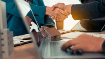 Close-up of a handshake between a real estate agent and a businessman with a small house plan in the background. Real estate, housing, transactions, contract concepts.